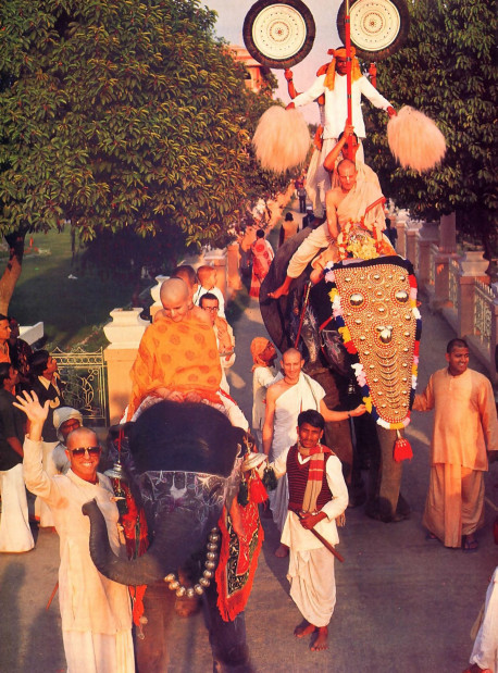 Atop an ornamented elephant, Parikajanghri steadies the Deities before him as Janannivasa leads the way in a colorful procession around the Mayapur temple gardens. A small elephant in the foreground joins Srila Bhavananda Goswami Visnupada, co-director of the Mayapur project and one of ISKCON's spiritual masters, in a wave to the camera.